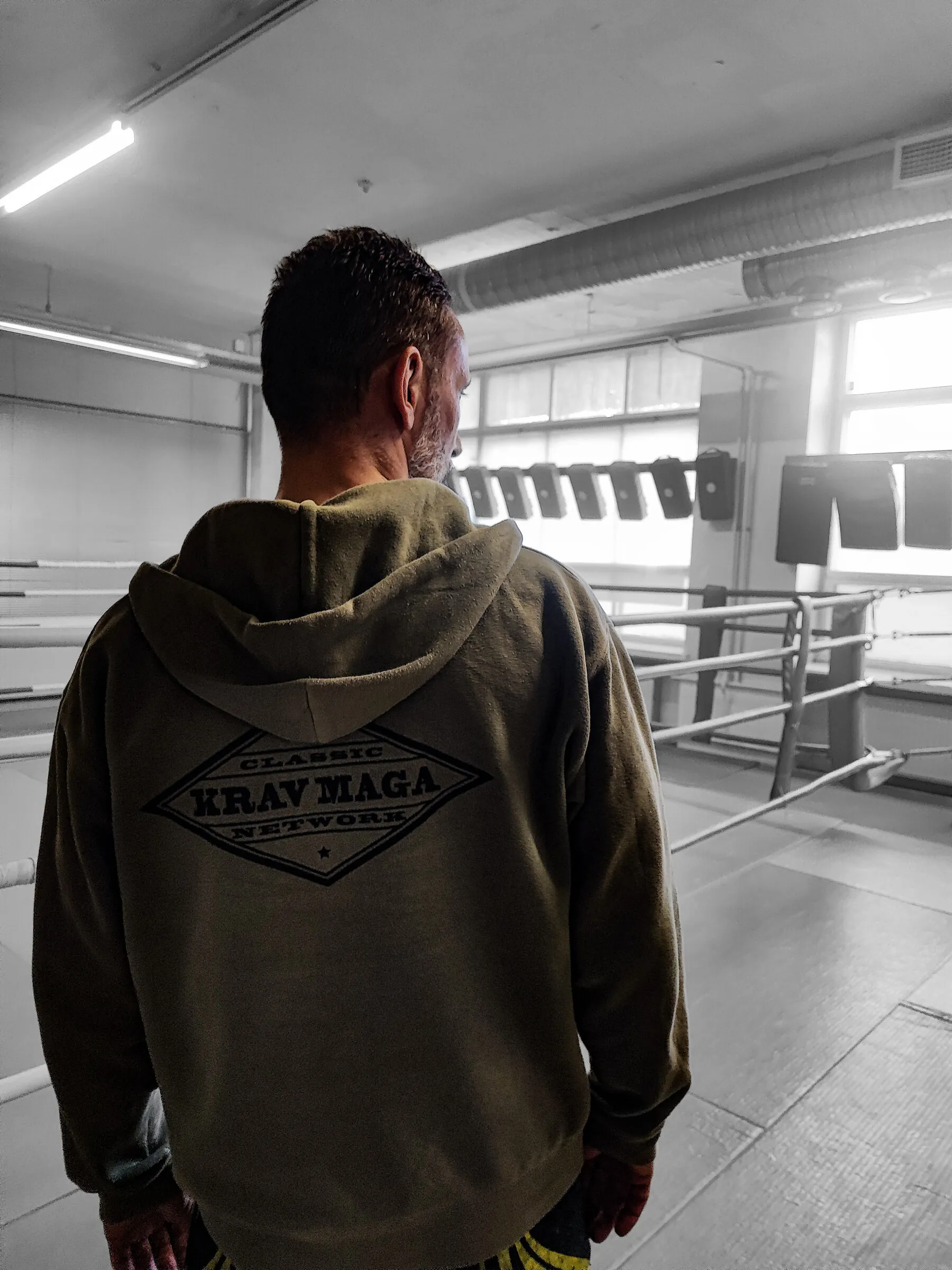 Ohjaaja takaapäin Classic Krav Maga Network -hupparissa, katse kohti harjoitussalia ja kehää