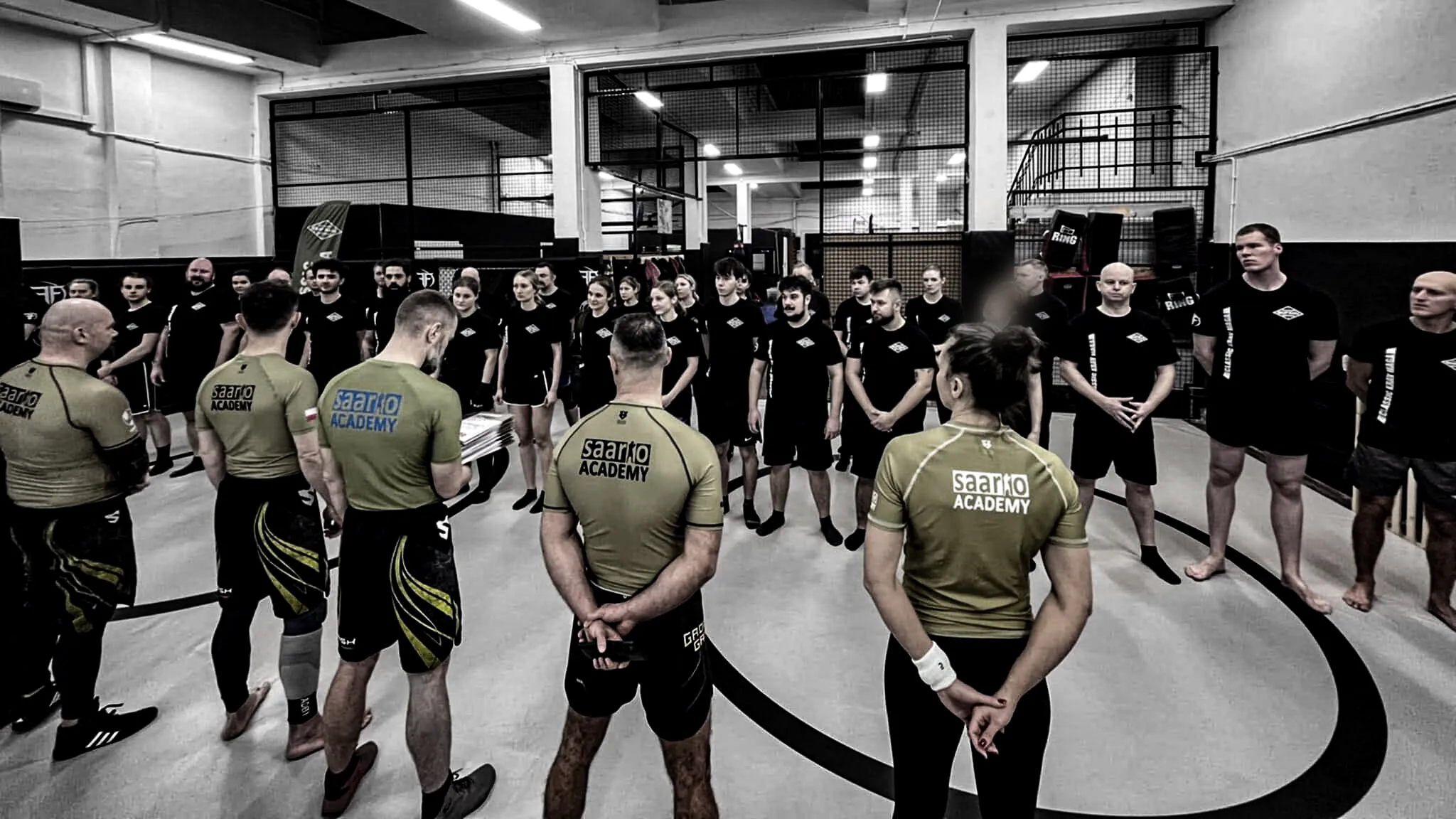 Grupa Saario Academy zebrana w kręgu wokół instruktora prowadzącego odprawę na macie treningowej